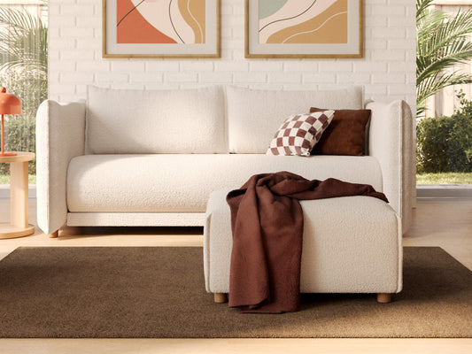 White sofa with checkered and brown cushions, matching ottoman with a brown throw, on a brown rug. Framed abstract art on white brick wall.
