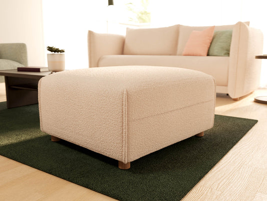 Cream fabric ottoman on dark green rug, near a light beige sofa with pastel cushions. Simple, modern living room setup with wooden floor.