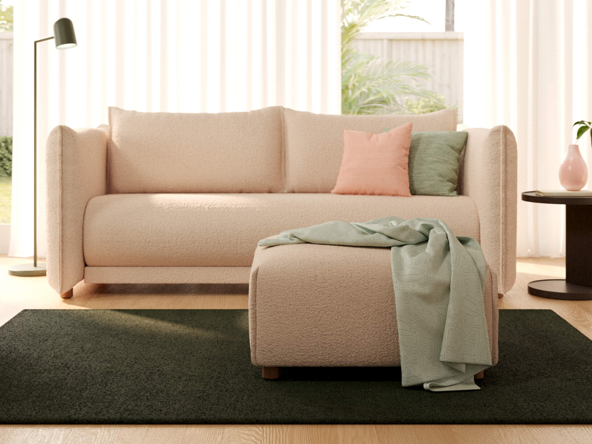 Cream sofa with pink and green cushions, matching ottoman draped with a green throw, on dark green rug. Round side table and floor lamp nearby.