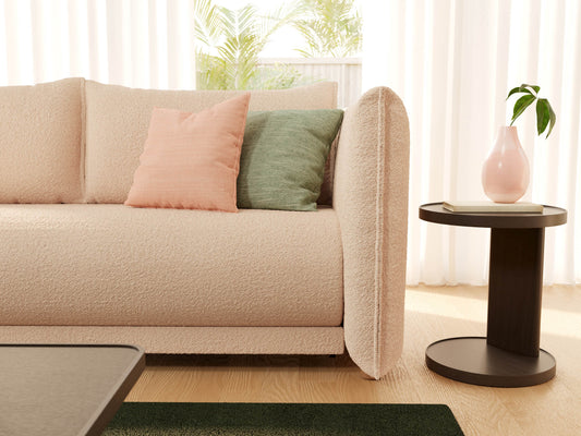 Cream sofa with peach and green cushions, beside a dark wood side table with a pink vase. Light curtains and green plants in the background.