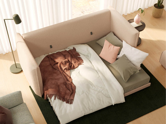 Beige corner sofa bed with white and green bedding, adorned with a brown throw blanket and multiple cushions, on a green rug.