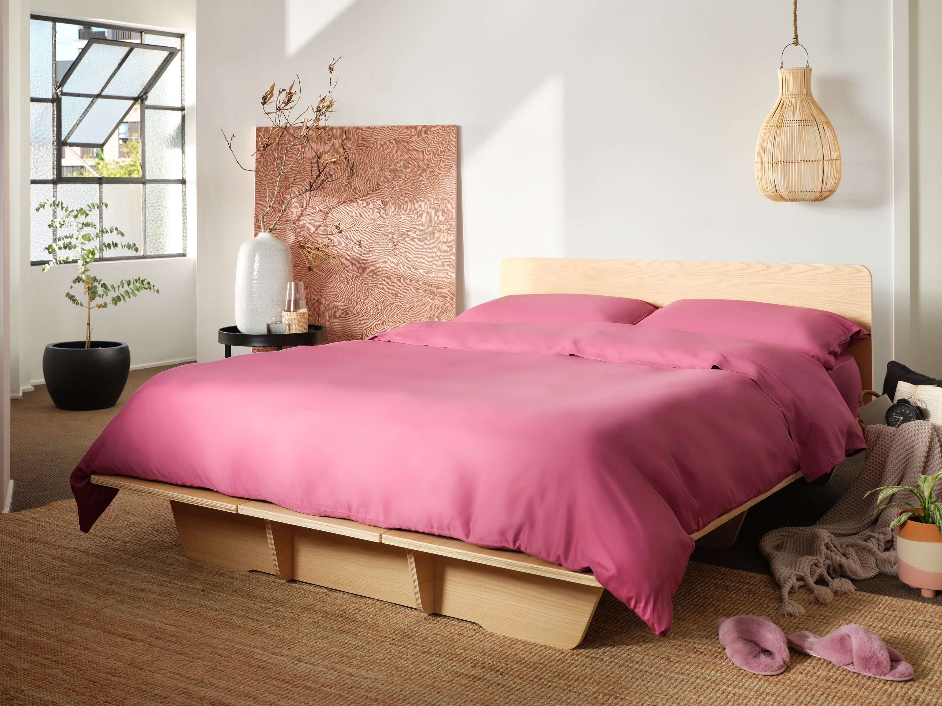 Bed with light wood frame and magenta bedding in a bright room. Natural decor includes a potted plant, woven light fixture, and wall art.
