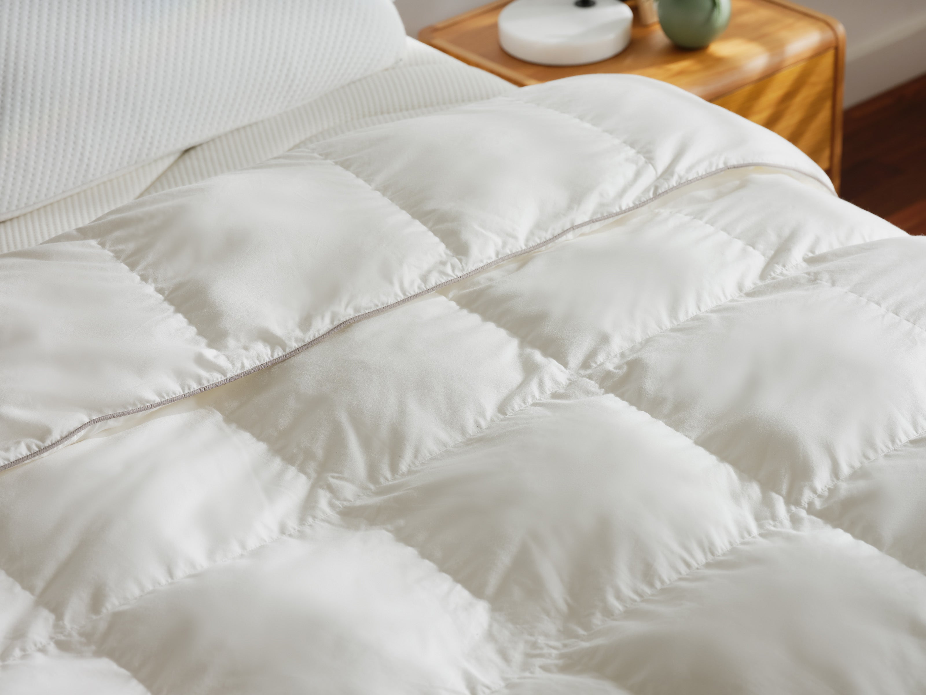 White quilted doona on a bed, with a light wooden bedside table holding a small plant and a round lamp.