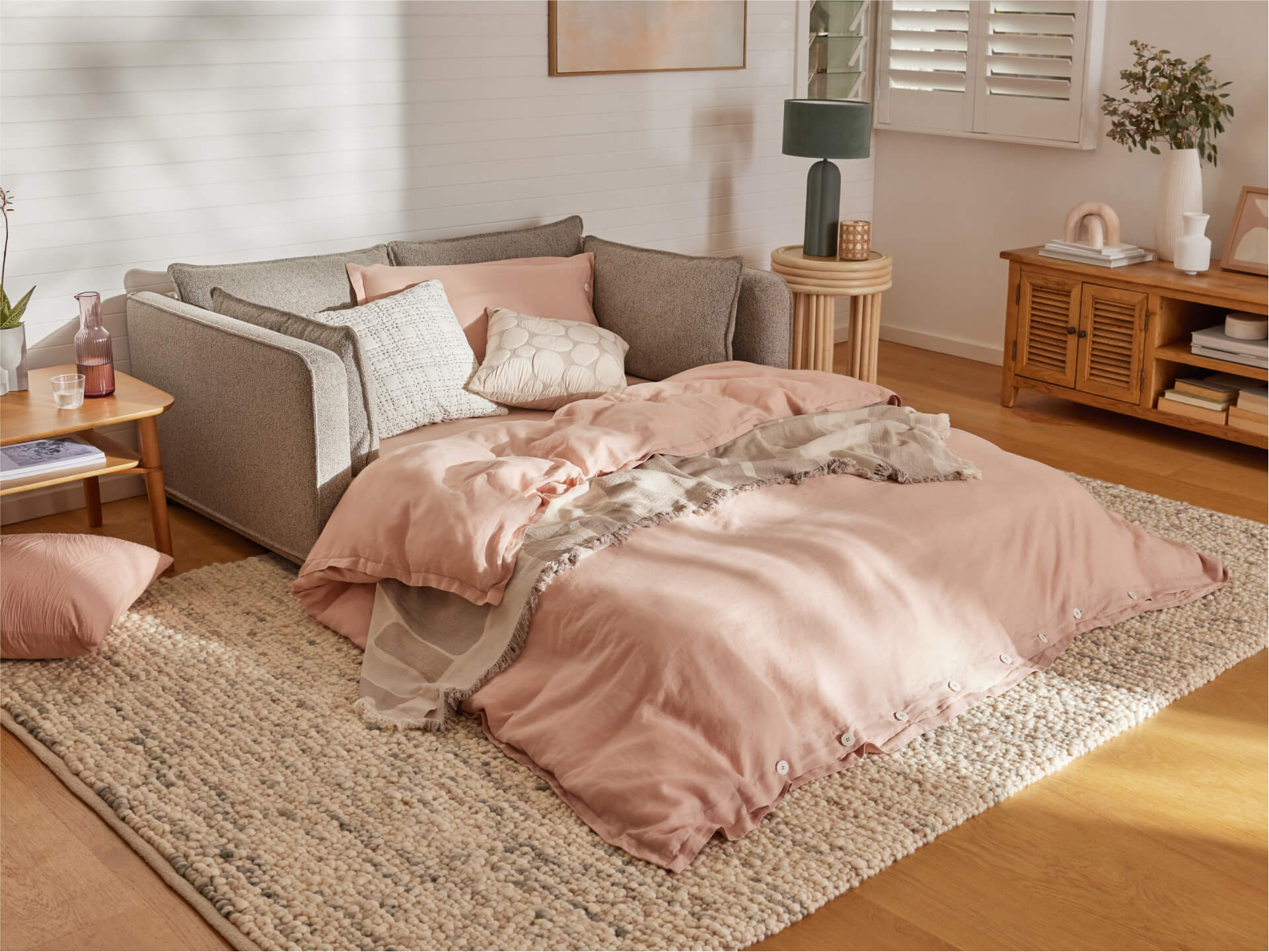 A cosy living room with a grey sofa bed, pink quilt, and cushions on a textured rug. Wooden table and cabinet with decor at the side.