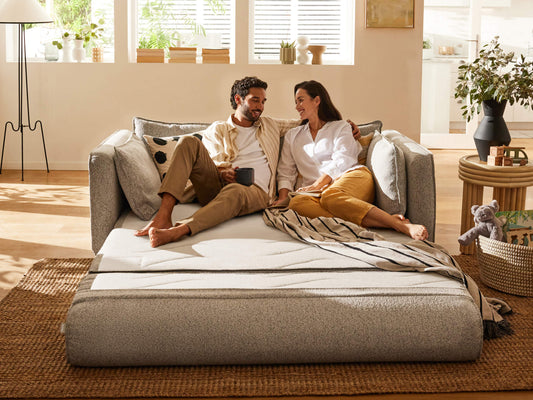 Couple lounging on a light grey sofa bed with cushions, surrounded by natural decor in a sunlit room, featuring a woven rug and indoor plants.
