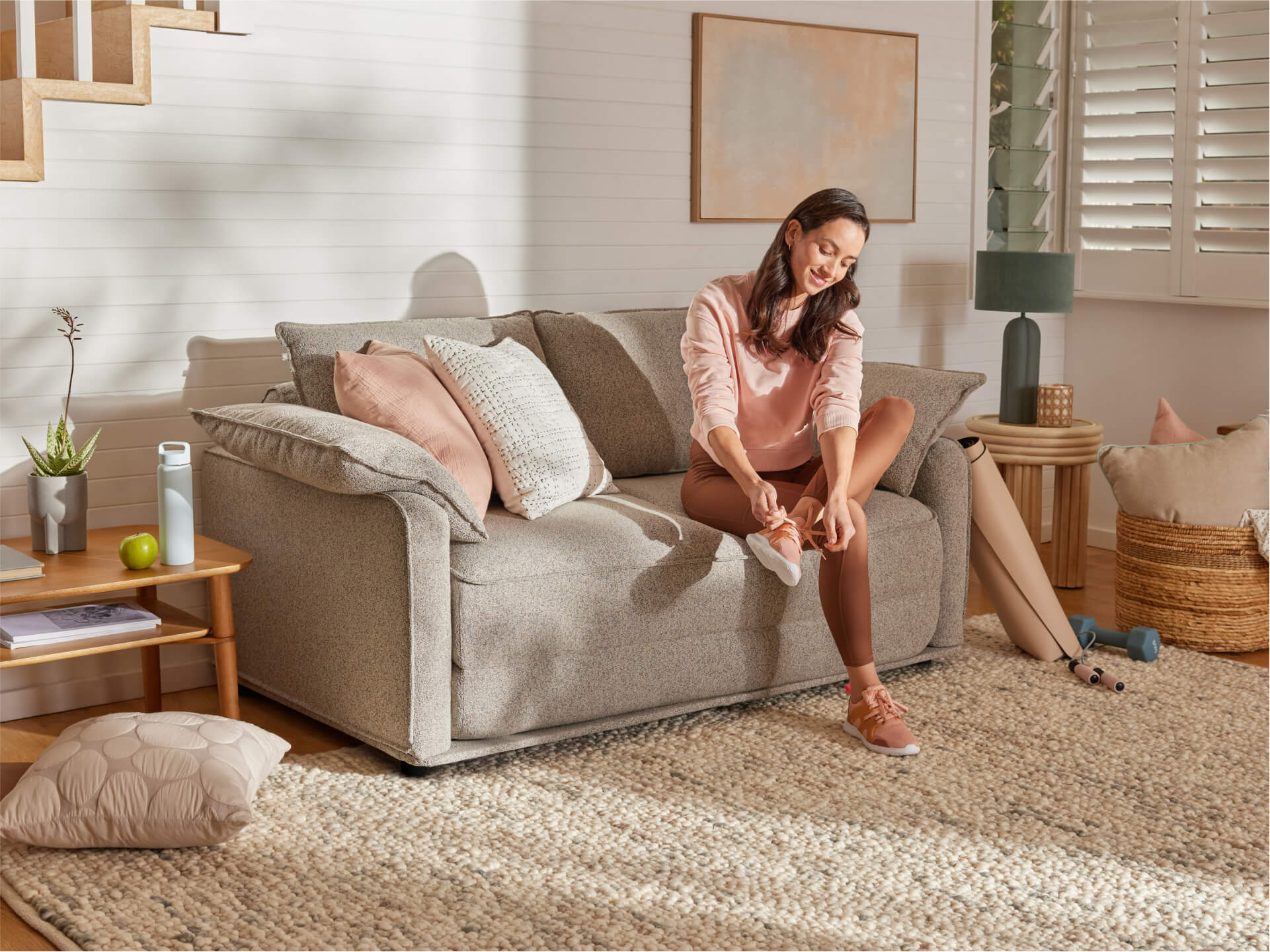 Woman in a pink top ties shoelaces on a light grey couch with cushions. Room features neutral decor and a cosy rug.