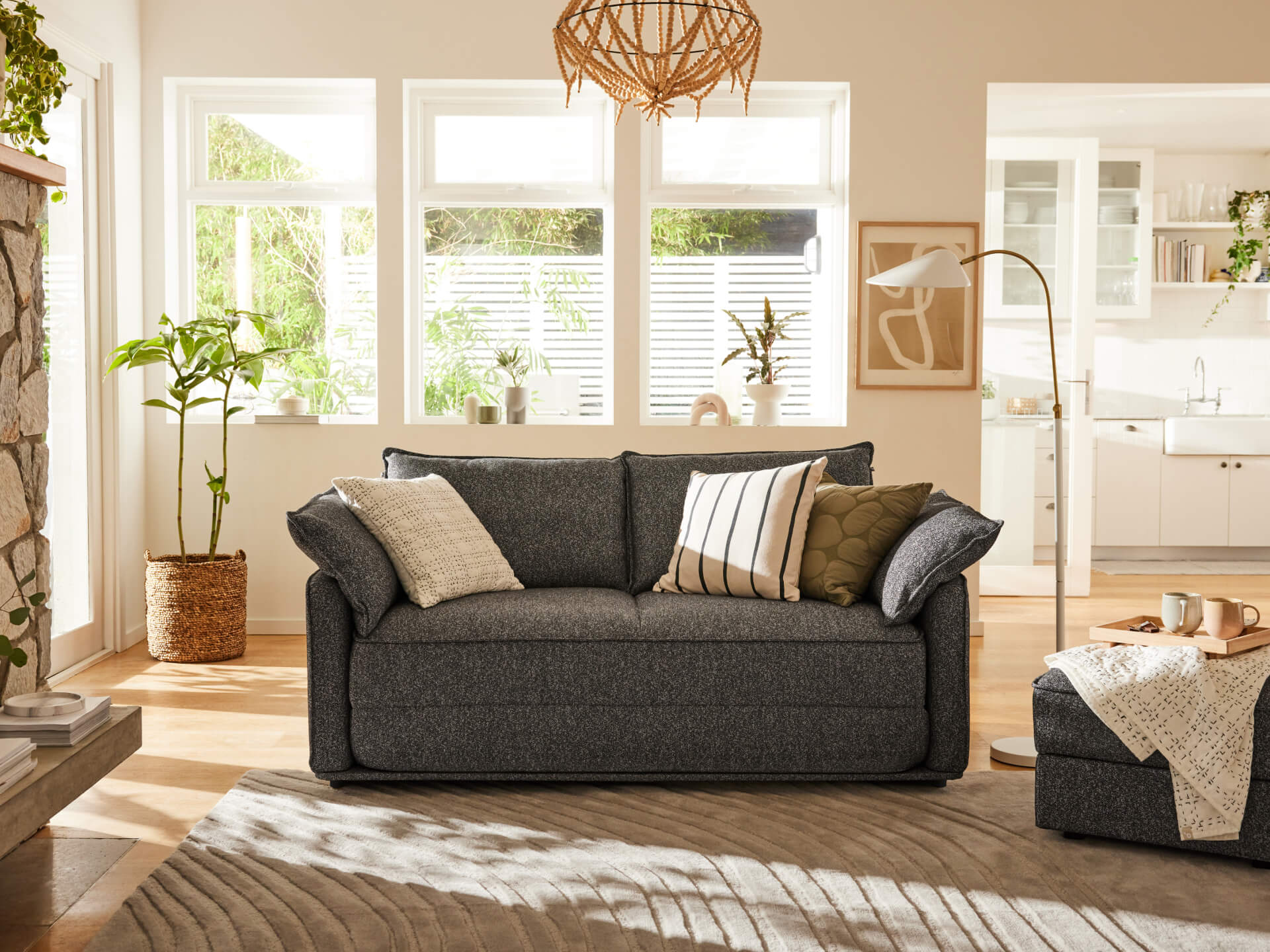 Cosy living room with a dark grey sofa adorned with various cushions. A floor lamp and indoor plants accentuate the sunny, neutral-toned space.