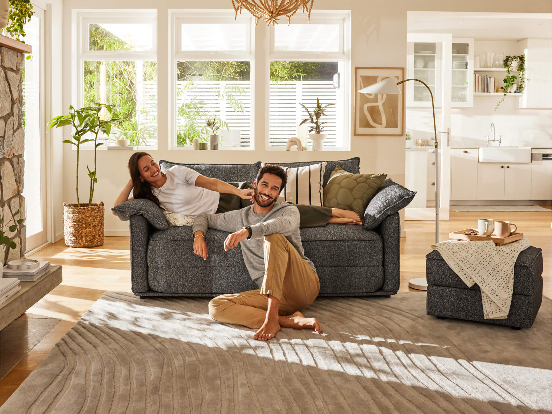 Couple relaxing on a dark grey sofa in a bright living room with plants, light walls, and a textured rug. Cosy, modern home setting.