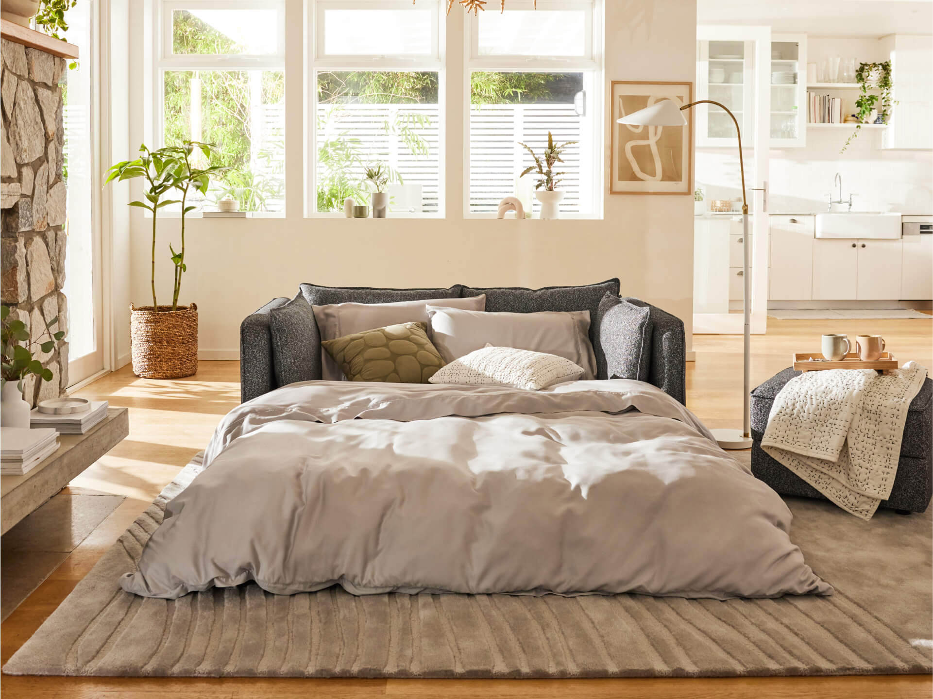 Stylish living room with dark grey sofa bed, light grey bedding, beige rug, and plants by a stone fireplace. Sunlit kitchen visible in the background.