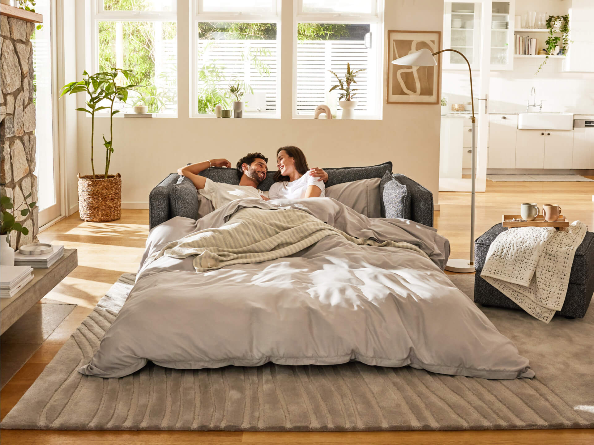 Couple lounging on a grey modular sofa bed with light grey bedding in a bright living room; plants and modern decor enhance the cosy atmosphere.