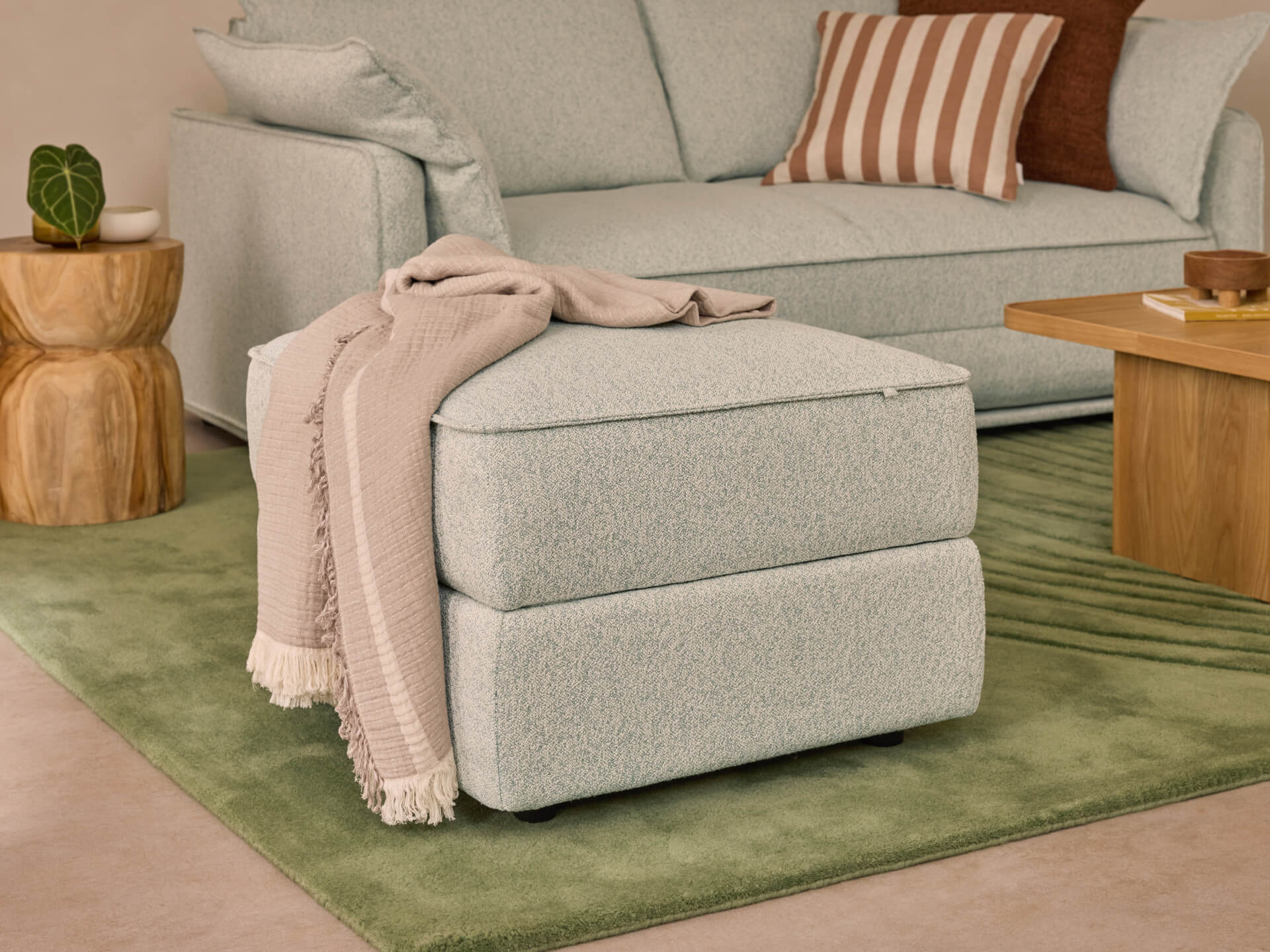 Light grey ottoman with a beige throw, placed on a green rug. Background includes a matching sofa with striped and brown cushions, and a wooden side table.