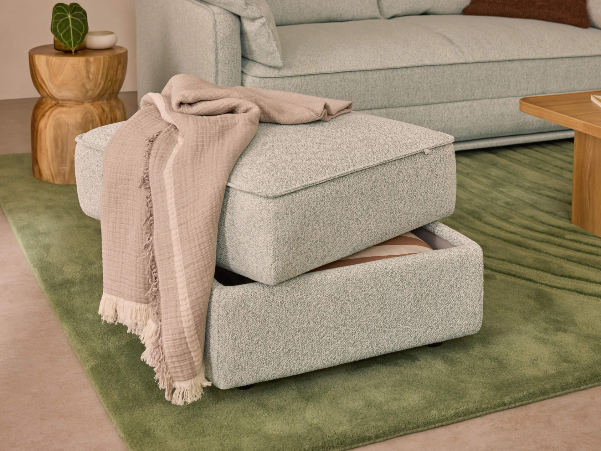 Light grey ottoman with storage on a green rug, topped with a beige throw blanket. Nearby are a wooden side table and a matching couch.