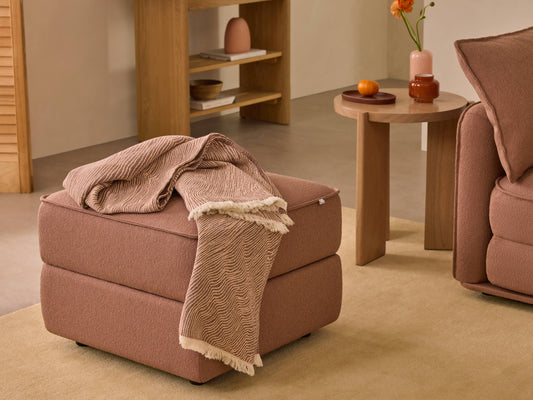 Brown ottoman with a patterned throw next to a matching sofa. Wooden side table with decor items in a cosy living room setting.