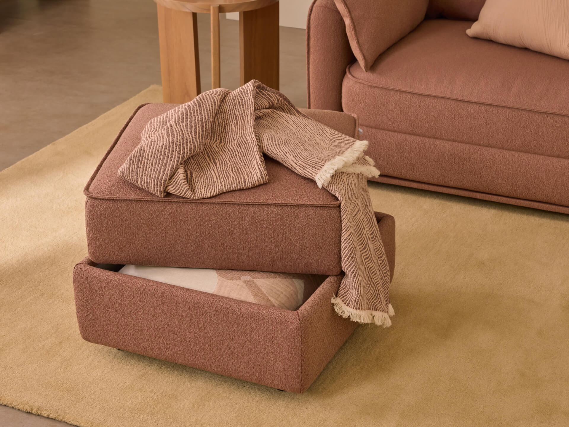 Pink fabric ottoman with storage, open to reveal a blanket inside. Matching lounge and pillow in the background on a beige carpet.