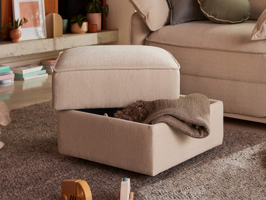 Cream ottoman with open lid revealing a blanket inside, on a grey rug. Beige sofa in the background with books and plants nearby.