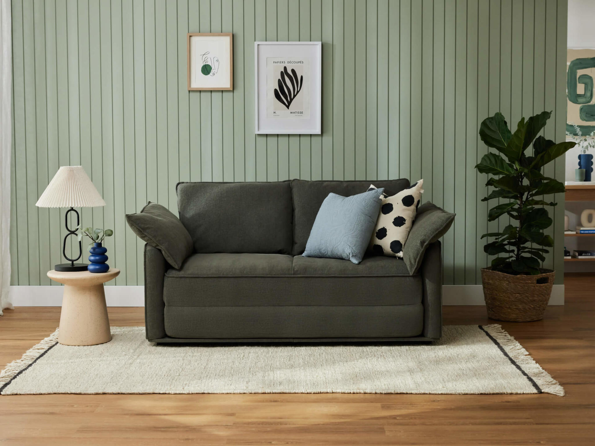 Dark green sofa with two cushions, against a light green panelled wall. Nearby are a small round table with a lamp and a potted plant on a rug.