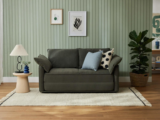 Dark green sofa with two cushions, against a light green panelled wall. Nearby are a small round table with a lamp and a potted plant on a rug.
