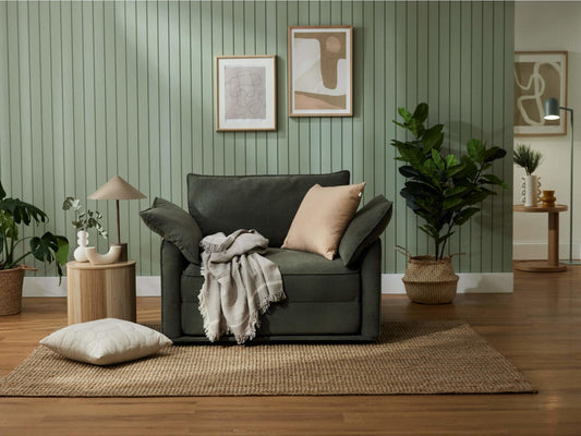 Green armchair with a beige cushion and throw on a woven rug. Surrounded by plants, side table, and framed art on green panelled wall.