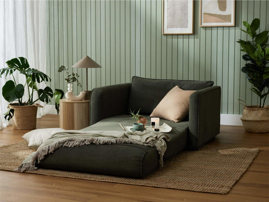 Dark green lounge chair on a textured rug with cushions, plants, and decor against a light green panelled wall in a cosy living room.