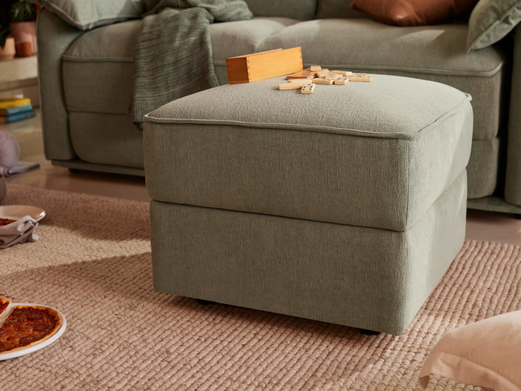 Light grey ottoman with a wooden game set on top, beside a matching couch. A textured rug covers the floor with a pizza plate nearby.