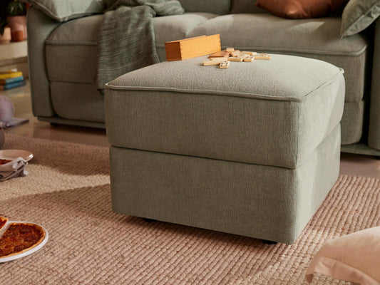 Light grey ottoman with a wooden game set on top, beside a matching couch. A textured rug covers the floor with a pizza plate nearby.