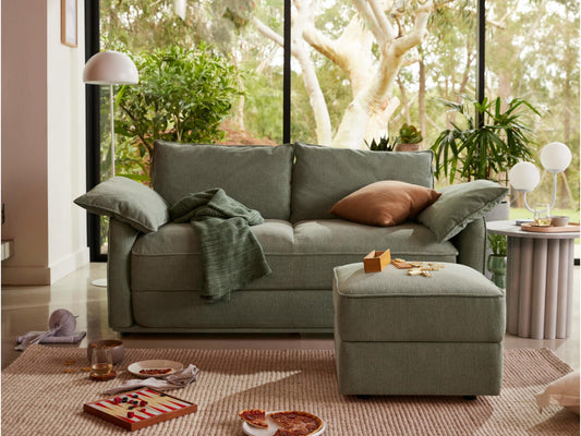 Cosy living room with a green sofa, matching ottoman, and indoor plants. Large windows reveal lush greenery. Neutral rug and small round table.