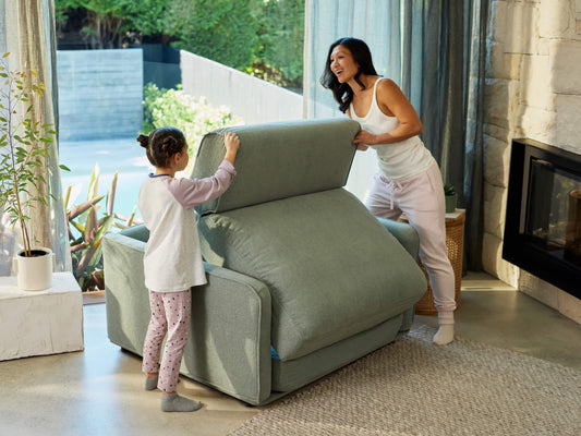 Woman and child assembling a light grey sofa in a bright living room with large windows and greenery outside.