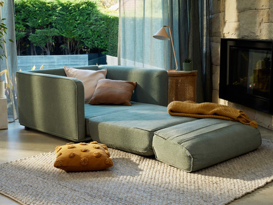 A green modular sofa bed with brown and beige cushions, a mustard throw, near a fireplace. It sits on a beige rug, with a wicker side table.