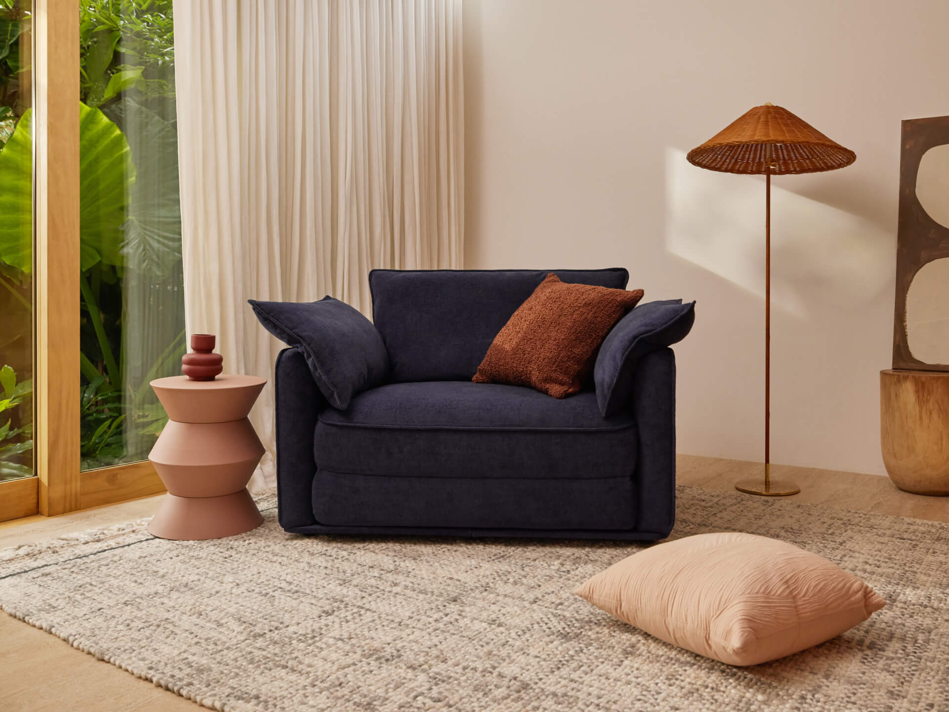 Navy armchair with brown cushion, geometric side table, rattan floor lamp, textured rug, and cream curtain. Tropical plants visible through window.