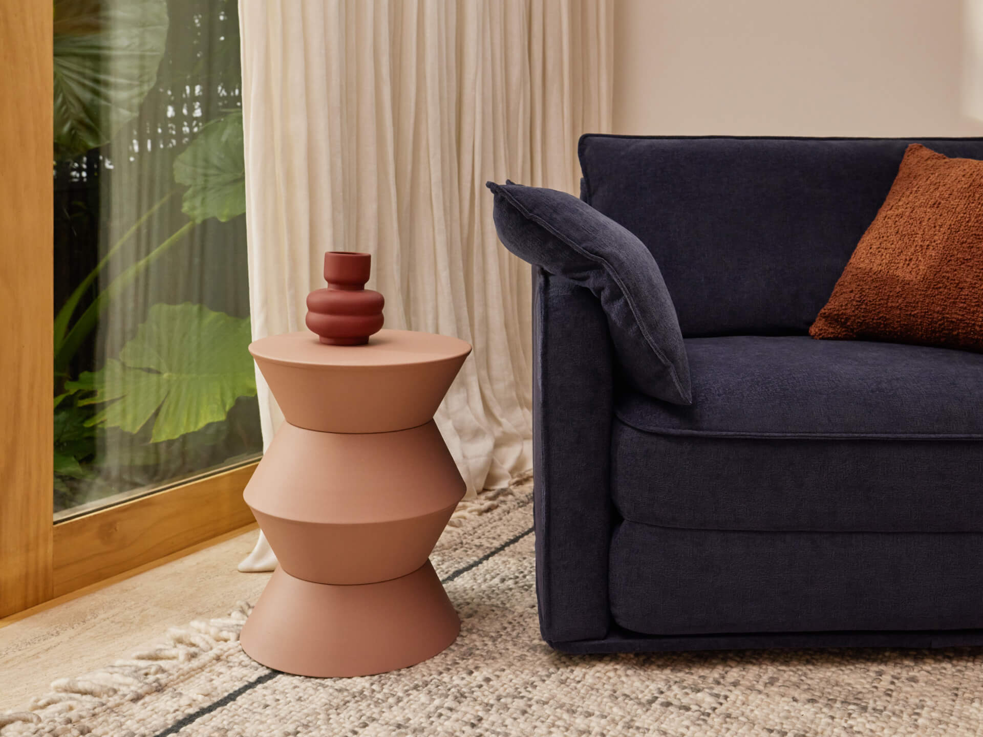 Navy blue sofa with a rust cushion, set next to a unique stacked terracotta side table. Sheer curtains and a large window in the background.