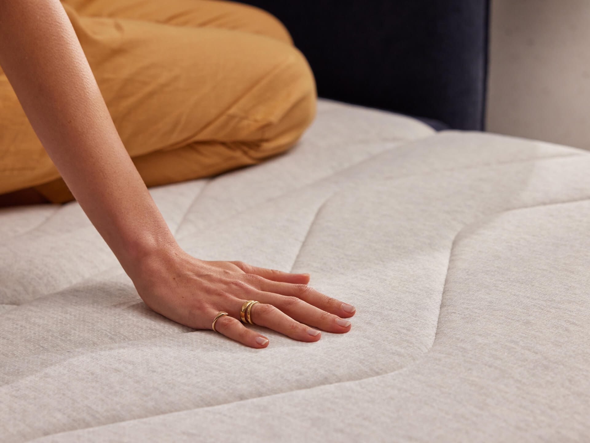 Hand pressing gently on a light grey mattress, showcasing its softness and texture. Person in brown pants partially visible.