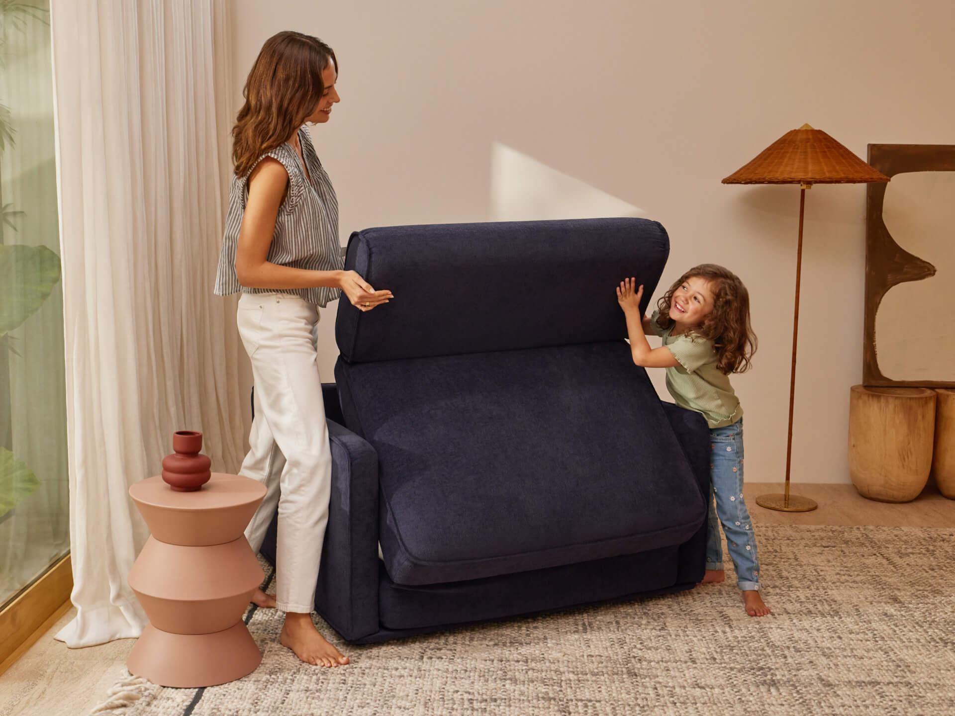 Woman and child adjusting a dark blue modular sofa in a living room with beige walls, patterned rug, decorative side table, and lamp.