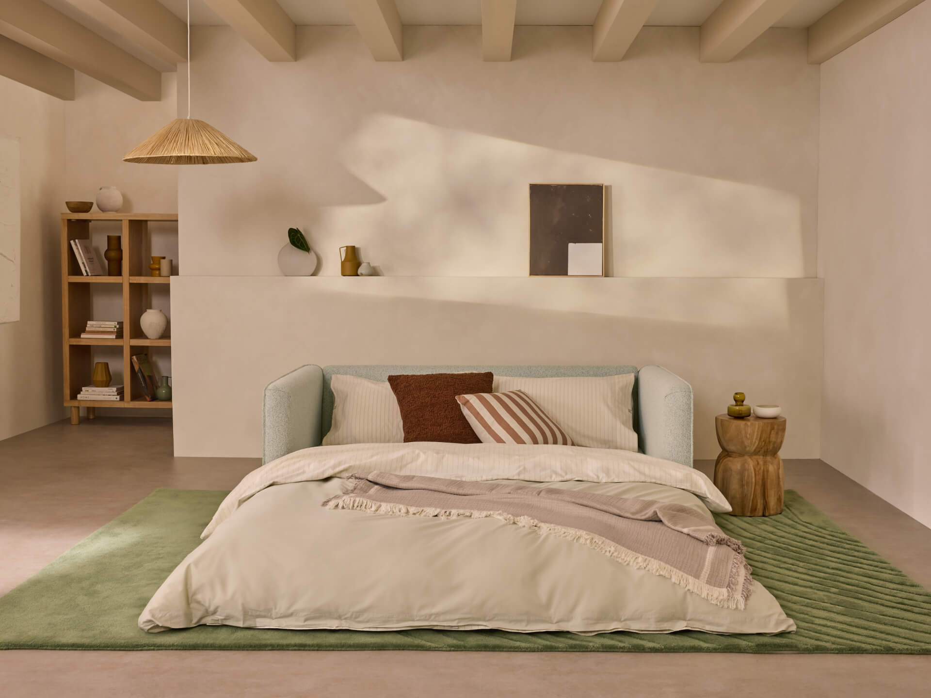 Minimalist bedroom with a light grey bed, brown and striped cushions, green rug, wooden side tables, pendant lamp, and a shelf with decor.