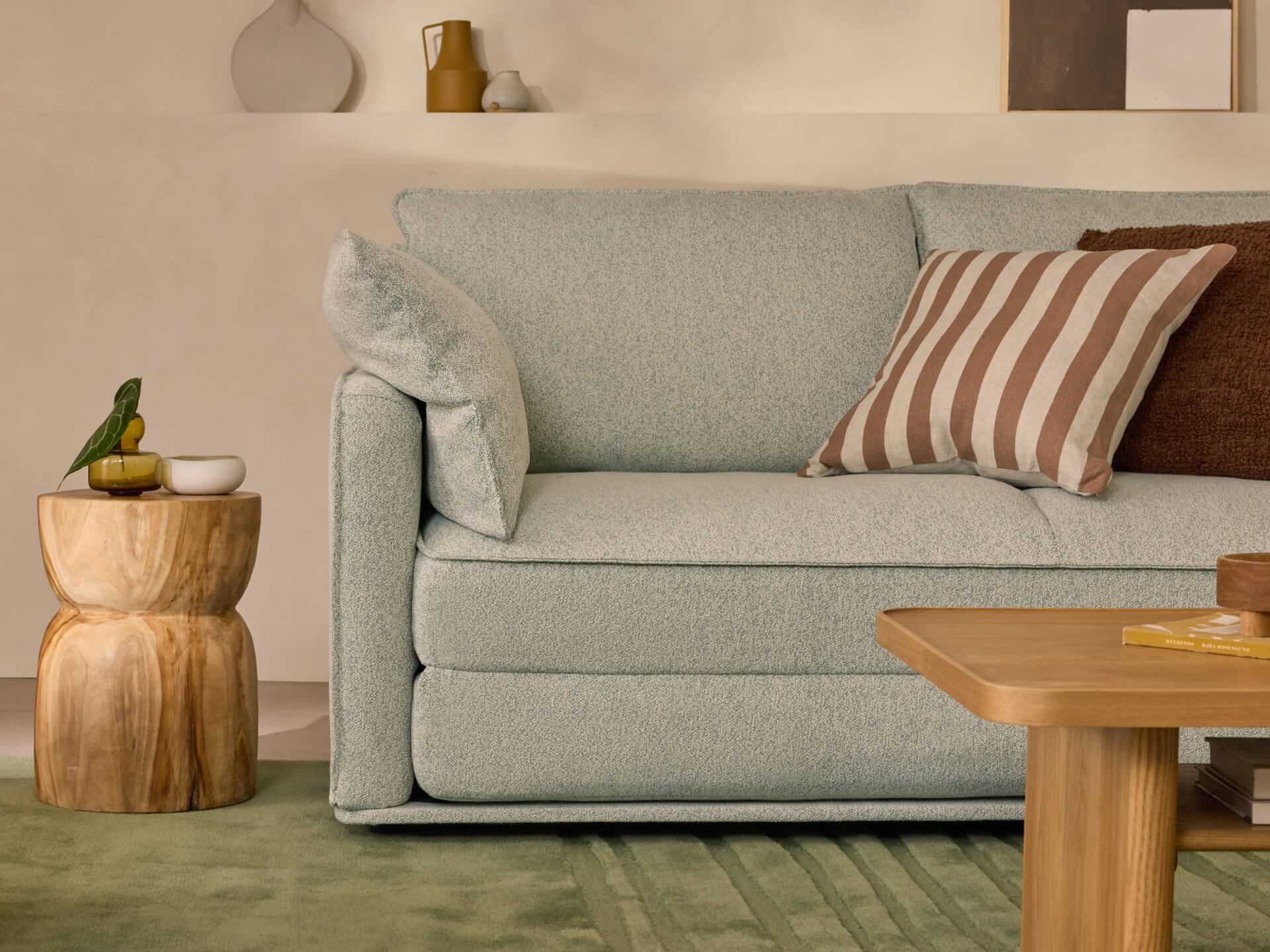 Light grey sofa with striped and brown cushions, beside a wooden side table with a small sculpture. Green rug and a wooden coffee table in front.