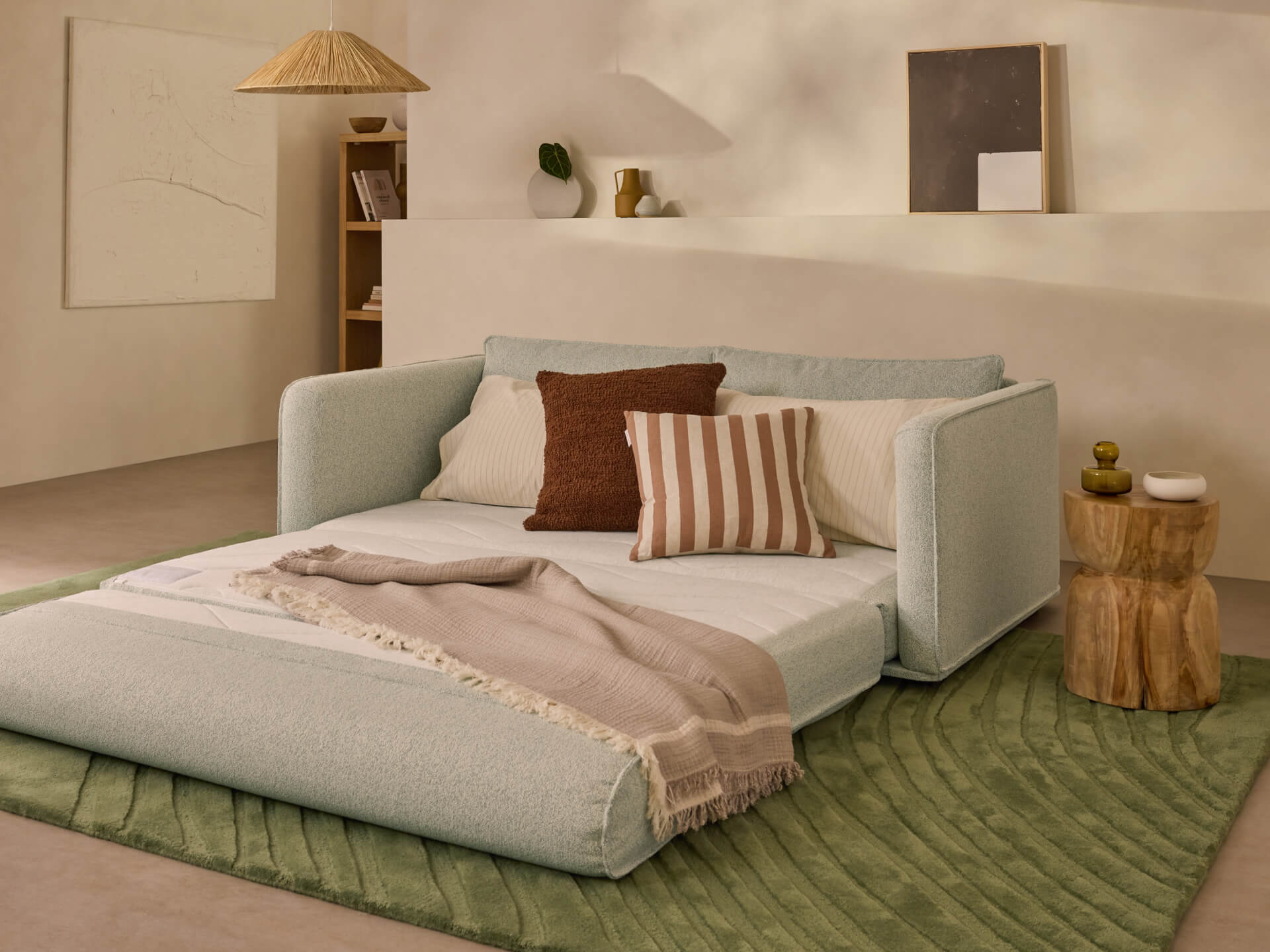 Sofa bed in light grey fabric extended on a textured green rug, adorned with a brown throw and striped cushions, next to a wooden side table.