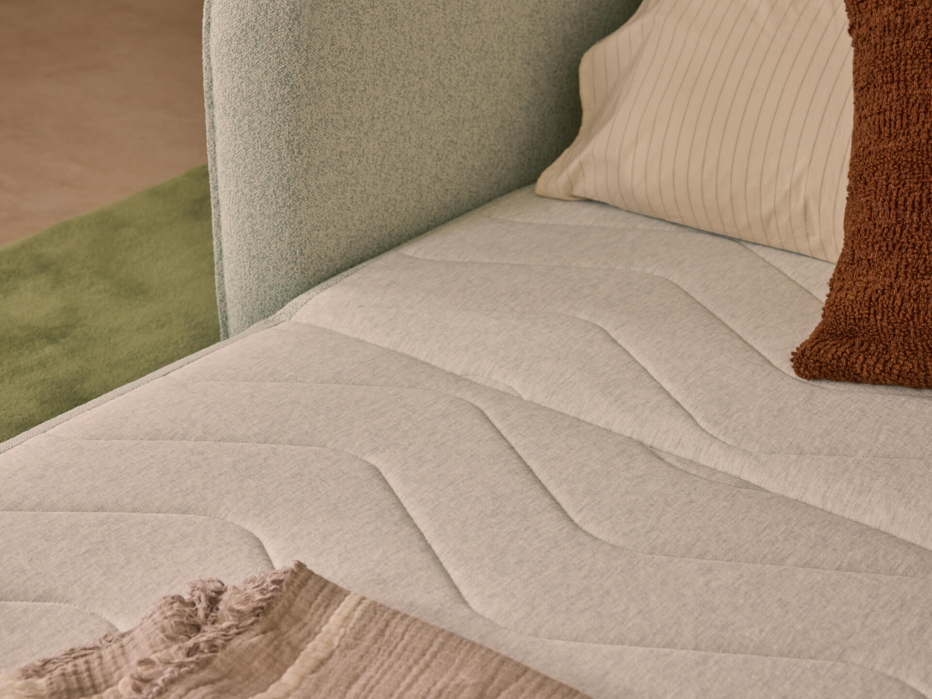 A plush light grey mattress with textured patterns, accompanied by brown and striped pillows. Green carpet partially visible in the background.