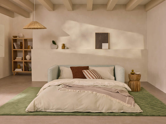 Minimalist bedroom with a light grey sofa bed, green rug, and neutral bedding. Wooden side table, wall shelf, pendant light, and beige walls.
