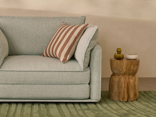 Light grey couch with a brown and white striped cushion, next to a wooden side table on a green textured rug.