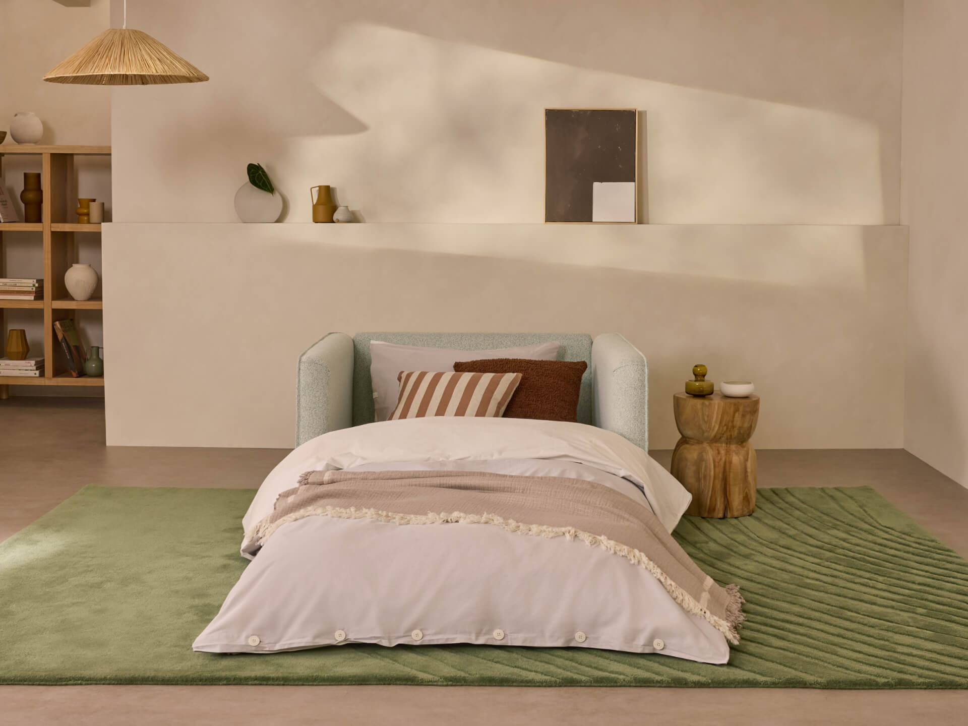 Minimalist bedroom with a light green sofa, white bedding on a green rug, and a wooden side table. Neutral decor accents the wall shelf.