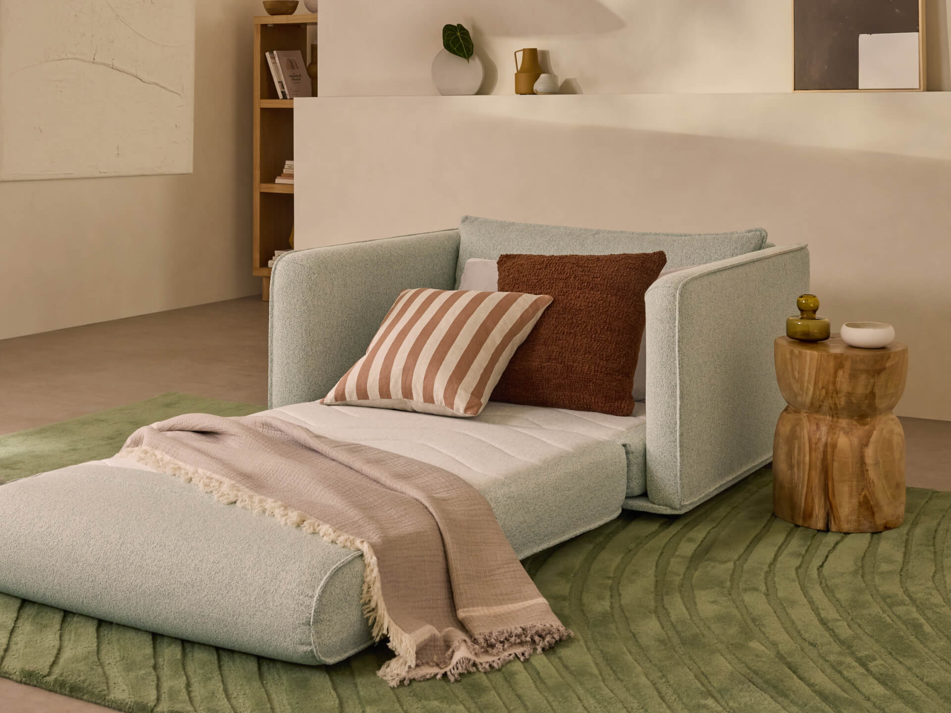 A light grey upholstered lounge chair with an extended ottoman, featuring striped and solid cushions, on a green rug next to a wooden side table.