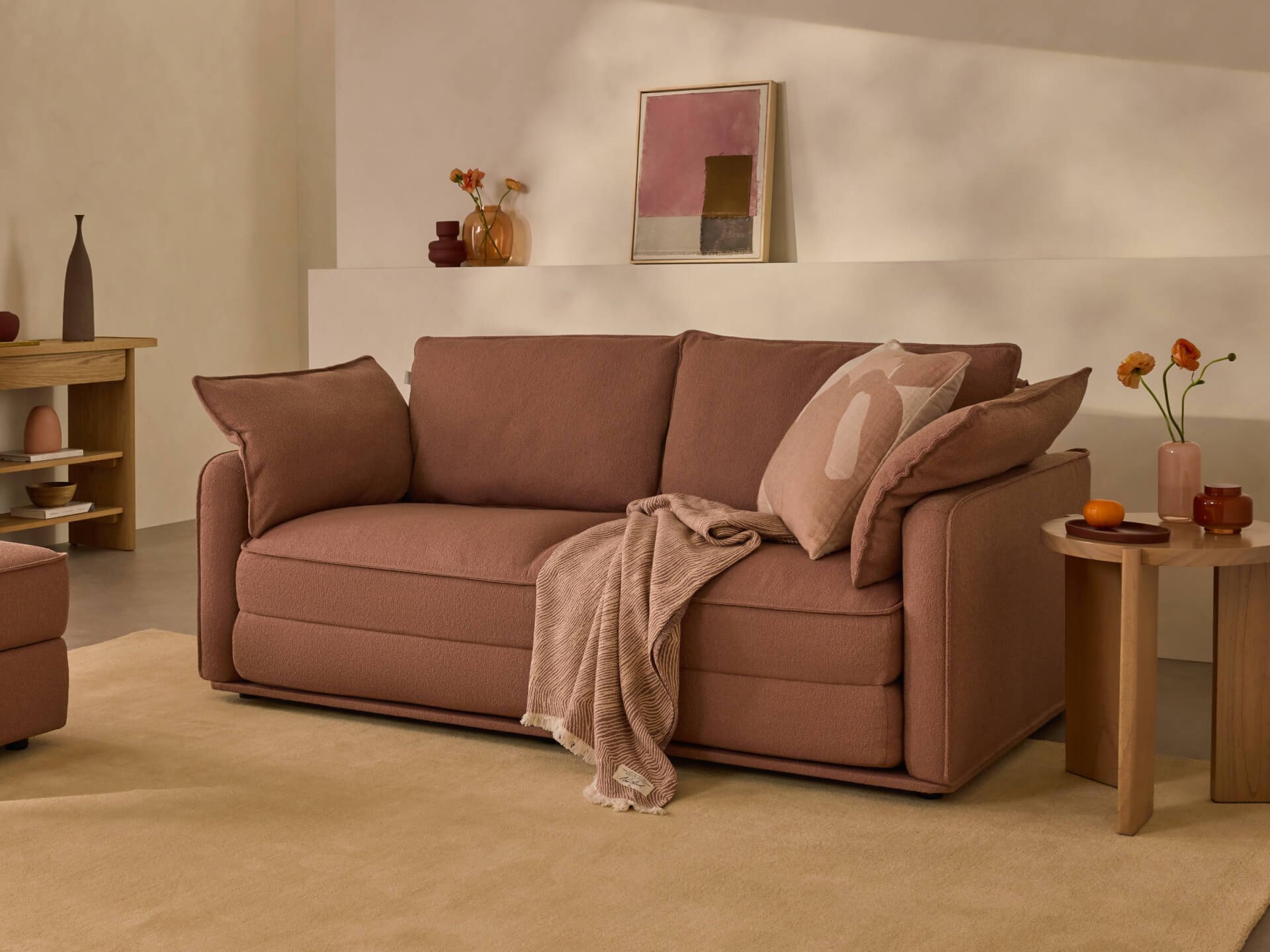 Cosy brown sofa with matching cushions and a beige throw, set in a warm, minimalist living room. Decor includes a side table and abstract artwork.