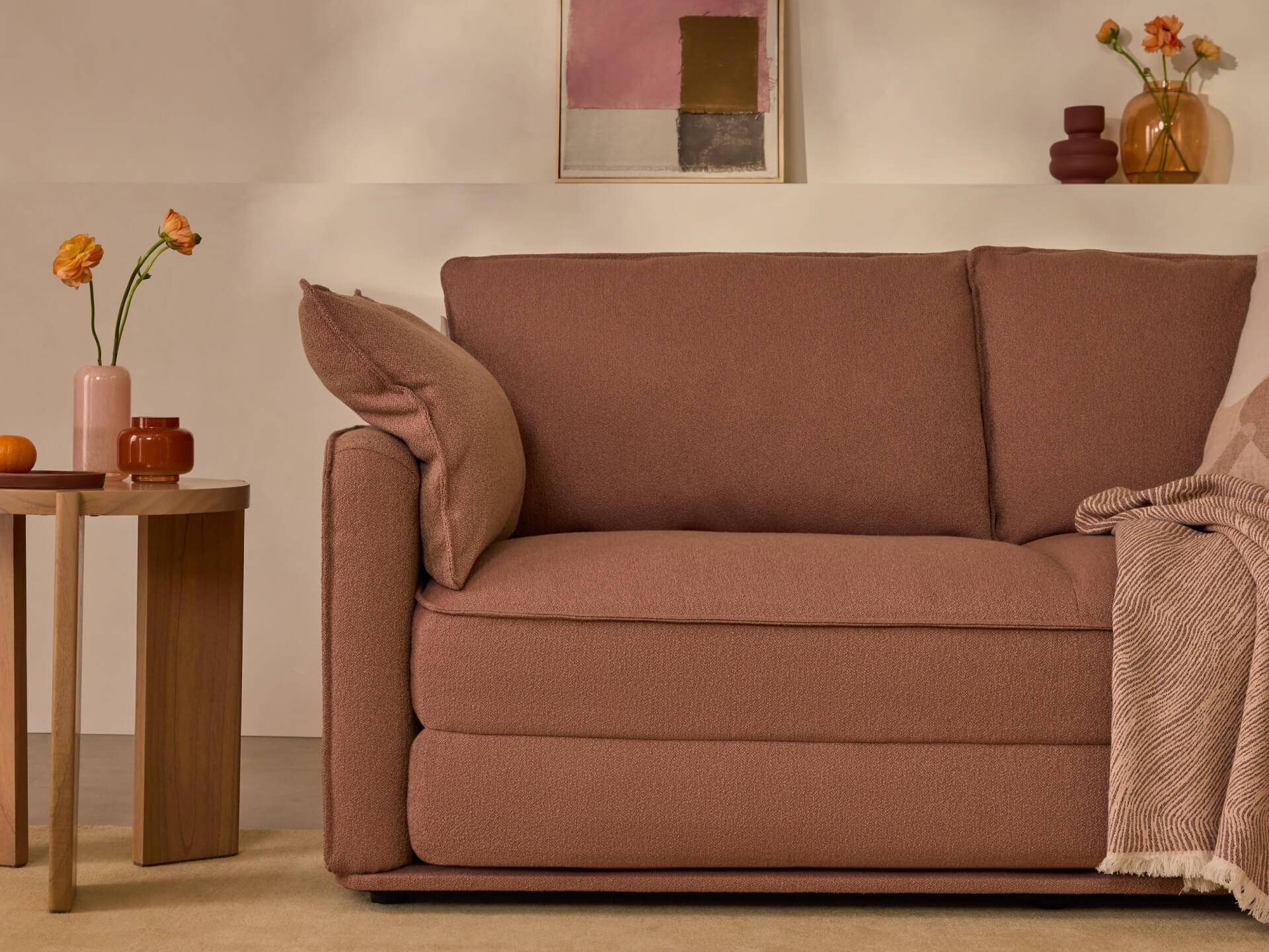 Brown loveseat with two cushions, a pink throw blanket, and side tables with vases, against a beige wall with abstract art.