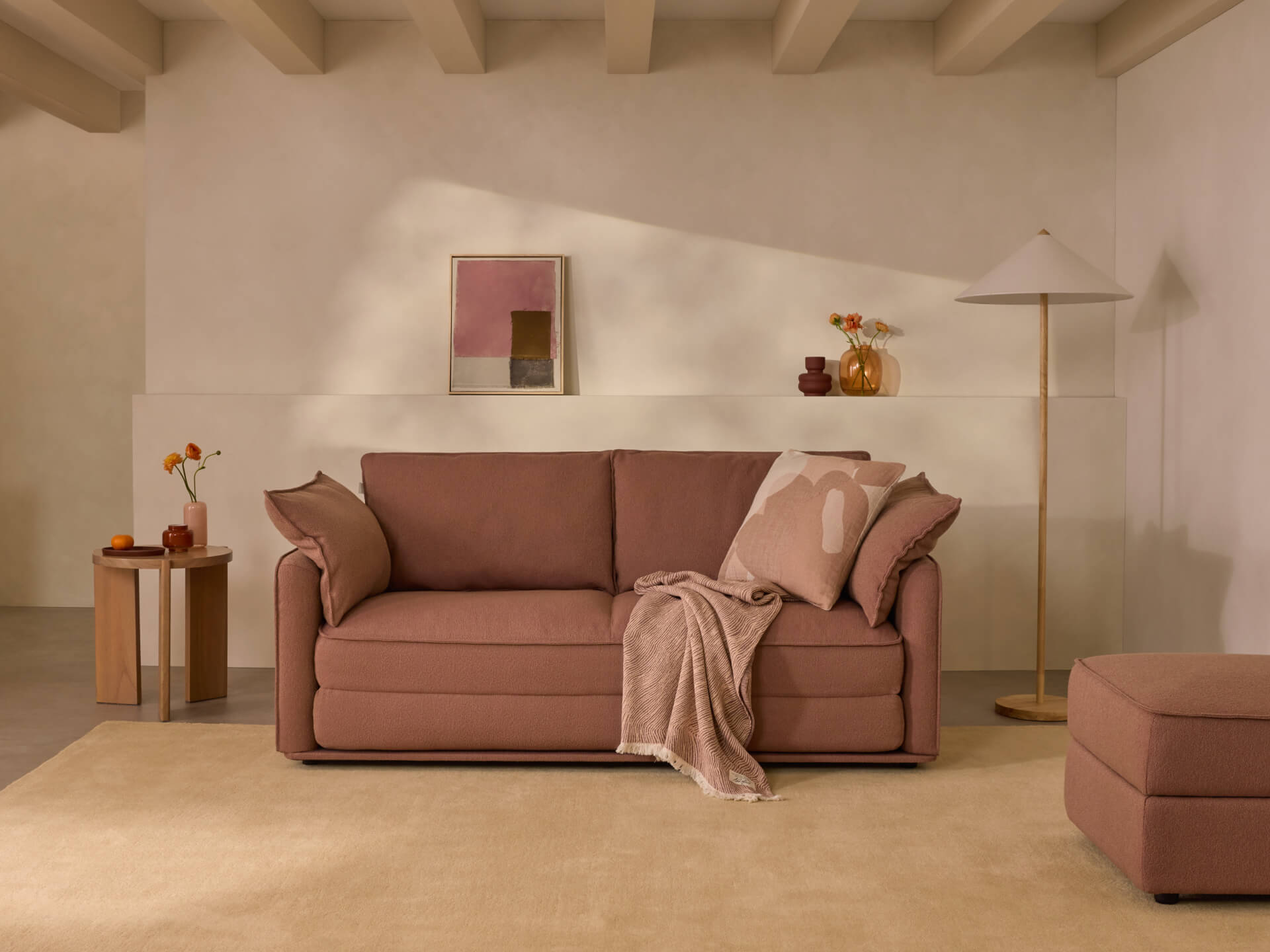 Modern living room with a brown sofa, matching ottoman, side table, beige carpet, framed art, and floor lamp. Decor includes vases and cushions.