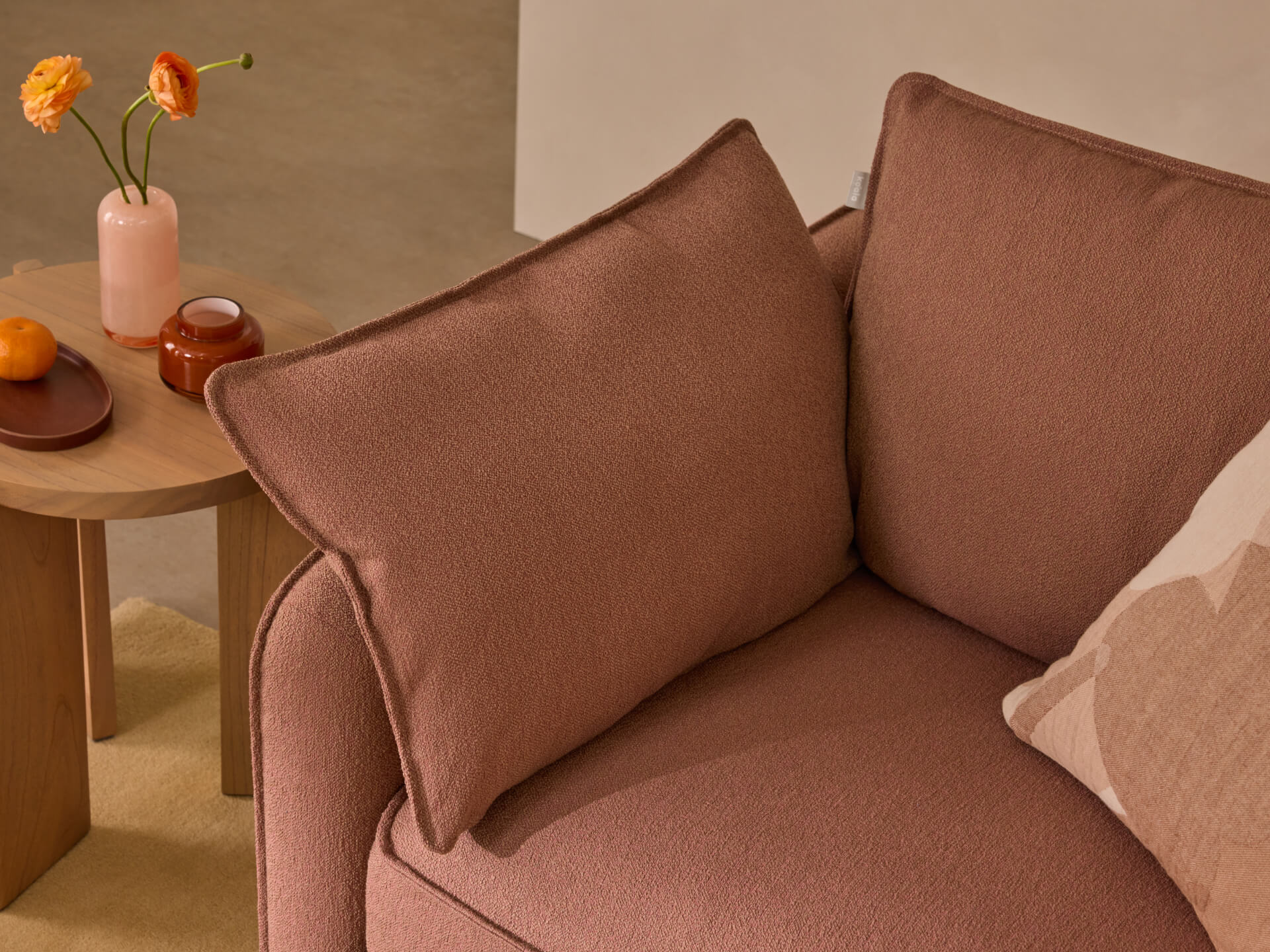 Brown fabric sofa with plush cushions, next to a wooden side table holding a vase with orange flowers and decorative items on a beige rug.