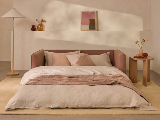 Cosy bedroom with a soft beige doona, pink sofa headboard, blonde wood side table, and minimalist wall art. Neutral tones and ambient lighting.
