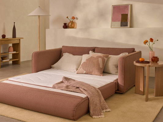 Modern living room with a pink sofa bed, beige and white cushions, wooden side tables, vases, and a floor lamp on a light beige rug.