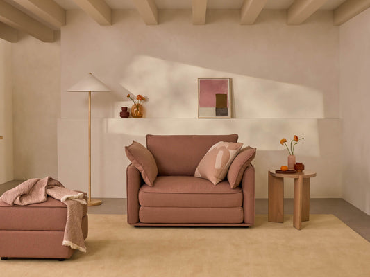 Warm-toned living room with a brown armchair, matching ottoman, light beige walls, minimalist artwork, and a wooden floor lamp.