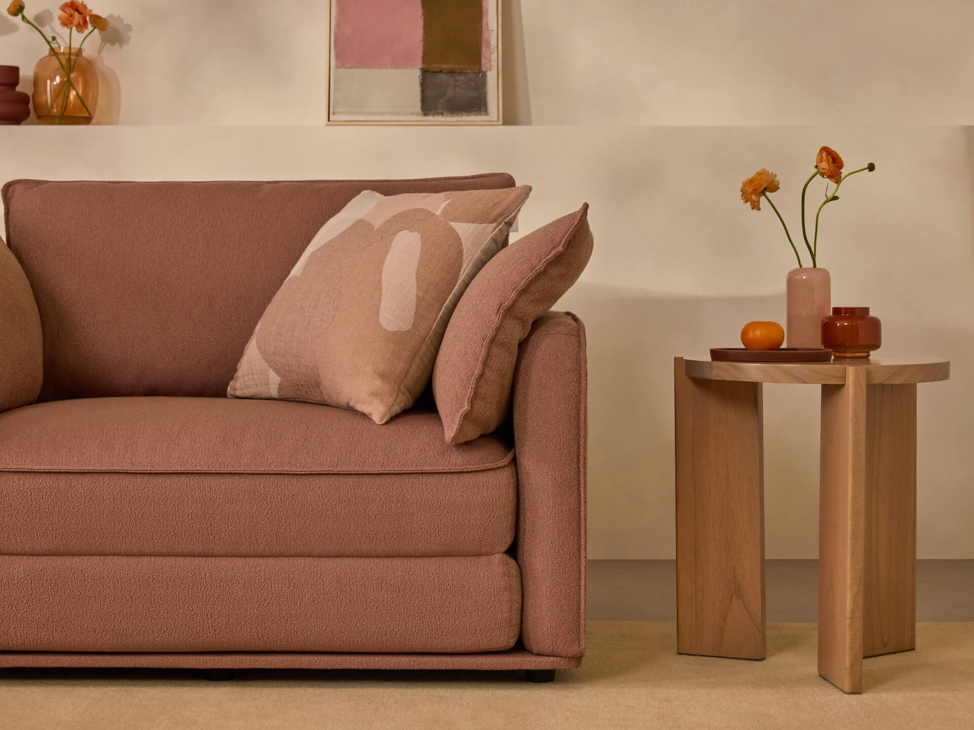 A brown sofa with patterned cushions next to a wooden side table. The table holds small vases with orange flowers. Minimalist decor setting.