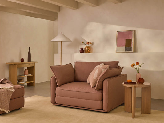 Modern living room with a large brown lounge chair, beige rug, wooden side table, and minimalist decor in warm tones, including vases and framed art.