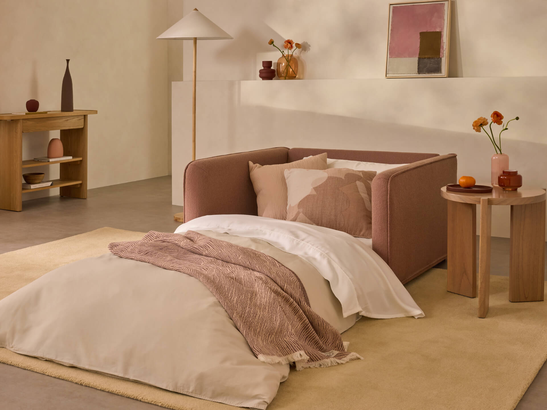 Modern room with a pink pull-out sofa bed, beige and pink bedding, wooden side table, and abstract art. Neutral tones and minimal decor.